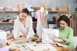 © JackF - Focused seamstress stitching linen fabric on sewing machine while female colleague drafting paper pattern at worktable, creating bespoke garment together in workshop