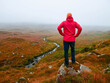 © mark_gusev - A man in a red jacket stands on a rock overlooking a river in Connemara, Ireland. The scene is serene and peaceful, with the man looking out over the landscape in a fog. Travel and tourism.