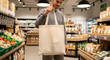 © BornHappy - A young man stands in a sustainable supermarket aisle holding up a plain beige reusable cotton shopping bag