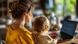 © VictorCanalesStudio - Mother multitasking on laptop with child by her side, balancing work and family life