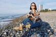 © SHOTPRIME STUDIO - Photographer woman sits on a pebble beach with a camera, wearing a plaid shirt and boots, capturing a coastal scene under bright sun, conveying relaxed outdoor lifestyle and creative travel moments