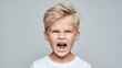 © MarkelovaVision - Angry young boy with blonde hair yelling with mouth wide open against a gray background.