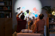 © DragonImages - Caucasian man, Caucasian woman, and young Caucasian girl sitting together creating animal shadow puppets on wall using colorful cutouts, engaging in playful family activity at home