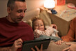 © DragonImages - Middle aged Caucasian man using digital tablet while lying in bed next to young Caucasian child smiling and relaxing, woman partially visible holding childs hand