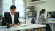 © Prathankarnpap - Two coworkers working independently in a modern office, one reviewing documents at a desk while another focuses on a computer