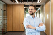 © Liubomir - Confident businessman in glasses and a light blue shirt stands with arms crossed, smiling in a bright modern office, conveying leadership, success and professional growth