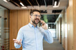 © Liubomir - Confident businessman engaging in a phone conversation, gesturing with an open hand while walking through a bright corporate office corridor, representing modern communication and networking