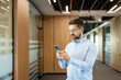 © Liubomir - Man in a light blue shirt and glasses actively checking his mobile phone, engaging with technology for communication and professional tasks while standing in a contemporary office building hallway
