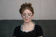 © Volha - Sad pensive young girl with collected hair and glasses looking down wearing a black dress with white lace collar sitting on a gray sofa, expressing melancholy and introspection