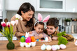 © Graphicroyalty - Mother and kids painting Easter eggs together. Creative family holiday tradition at home. Smiling woman with children decorating colorful eggs with brushes in the kitchen for Easter celebration.