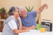 © luciano - Happy elderly couple sitting together at a table in the garden take selfies with smartphone while enjoying free time at home. Authentic retirement lifestyle for two modern smiling seniors