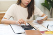 © KMPZZZ - Stressed asian young business woman, employee using calculator to calculate expenses of monthly, hand holding bills and receipt for to payment on table at home. Financial, finance of banking concept.