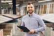 © anatoliycherkas - Retail manager smiling, holding clipboard, standing in a large hardware and flooring store, checking inventory