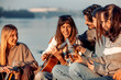 © ivanko80 - Friends enjoy a sunny day by the river while one plays guitar and others share drinks and laughter