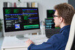 © Mediaphotos - Young man sitting at desk working on computer with multiple monitors displaying programming code, wearing glasses, focusing on screen in office setting