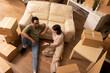 © DC Studio - Multiethnic couple sits indoors with cardboard boxes, discussing apartment arrangements. Diverse spouses relaxes on floor, leaning against couch, enjoying break during moving day in new home.