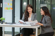 © aekachai - Two professional Asian women collaborating at a white desk in a modern office setting. One woman is using a calculator while the other reviews a document intently. They are working together