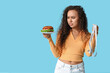 © Pixel-Shot - Thoughtful young African-American woman with measuring tape looking at burger on blue background. Diet concept