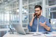 © Liubomir - Young man in casual clothes communicating on smartphone, sitting at desk in creative office, discussing important project or making business decisions with a focused expression
