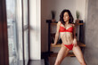 © Vlad - Seductive pretty young woman wearing lace red lingerie, posing looking out the window at her apartment. Copy space. Erotic, seduction concept
