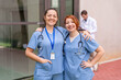© StockMe - Two happy female nurses in scrubs smiling, standing outside hospital building, with a colleague in background