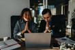 © qunica.com - Two professionals sit side by side, hands pressed together in a praying gesture while looking at a laptop. A modern office setting with documents, a cup, and a Christmas tree in the background.