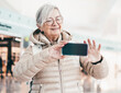 © luciano - Smiling senior caucasian traveler woman with backpack at the airport gate waiting for departure flight, elderly woman using phone