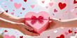 © Lumora Studio  - Close-up hands of a couple holding a heart-shaped pink gift box with ribbon against a background of floating hearts, Valentine's Day celebration, romantic surprise, love and anniversary concept