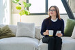 © Valerii Honcharuk - Relaxed mature woman with smartphone cup of drink sitting on sofa at home