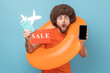 © khosrork - Surprised man with Afro hairstyle standing with orange rubber ring, holding passport document, sale card and showing cell phone with empty display. Indoor studio shot isolated on blue background