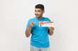 © khosrork - Portrait of attractive cheerful happy man wearing T- shirt standing with gift voucher and pointing at camera, certificate coupon for you. Indoor studio shot isolated on gray background.