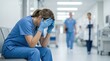 © Mariya Sorvacheva - Female healthcare worker in blue scrubs sitting on a bench in a hospital corridor, holding her head in distress with medical staff visible in the background