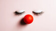 © Bonsales - False eyelashes and a red clown nose forming a minimalist, humorous face on a pastel pink background, representing beauty and playfulness