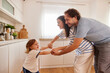 © Stockphotodirectors - A joyful family plays together in their kitchen during a sunny afternoon. The parents hold hands with their child, creating a fun and engaging moment filled with laughter and love.