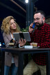 © Nikola Spasenoski - Casual evening meeting at cafe with a woman and man discussing over a tablet and coffee, sharing ideas and having a phone conversation, fostering a collaborative and friendly atmosphere.