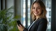 © Zabi - A radiant young businesswoman with long wavy hair smiles confidently at the camera while holding a smartphone in an elegant modern office with large windows behind her.