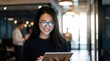 © CuteBee - A cheerful young Asian woman wearing glasses smiles while confidently using a digital tablet in a brightly lit modern office hallway.
