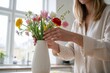 © KraPhoto - Frau arrangiert bunten Frühlingsstrauß mit Tulpen und Ranunkeln in weißer Vase am Fenster