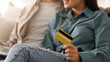 © Prostock-studio - A young couple sits together on a couch in a cozy living room. One person holds a yellow credit card, while browsing online shopping on a laptop nearby.