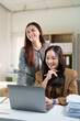 © MINAE - Two smiling Asian business women collaborating and discussing work, looking at a laptop computer while sharing ideas and ensuring productivity in a modern office environment