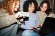 © Studio Marmellata - Three young women with curly hair are gathered around a laptop, one holding a card, smiling and engaged in online activity.