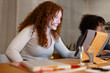 © Studio Marmellata - A young woman with curly red hair smiles while looking at a tablet. Another person is visible in the background, also working on a device.