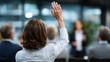 © Maksym - Clean, modern stock-style image of a businesswoman seated in a conference room raising her hand, colleagues attentive around her, speaker presenting in the background, shallow dept