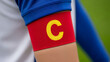 © Saieem - Close-up on a soccer player's arm wearing a red captain's armband with a yellow 'C' emblem, symbolizing leadership and team spirit.