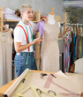 © JackF - Tailor stands in the workshop near a mannequin with a measuring tape. Male fashion designer is working on a collection of clothes and stands against the background of shelves and racks with fabrics