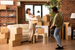 © DC Studio - Young diverse couple standing in modern living room, carrying cardboard box and potted plant during apartment relocation. Happy multiethnic partners moving belongings in new home together.