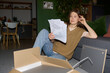 © pressmaster - Caucasian young adult woman sitting on floor assembling furniture, holding instruction sheets and examining parts, cardboard box and tools scattered nearby in modern living room