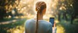 © mobile - back view of a young woman with a long braid holding a smartphone on a sunlit park path with warm golden bokeh, peaceful and contemplative mood