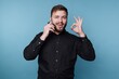 © bodnarphoto - Joyful young man on the phone sharing good news against a vibrant blue backdrop while making an okay hand sign