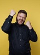 © bodnarphoto - Excitement and joy expressed by a man in a black shirt against a bright yellow backdrop during a moment of triumph